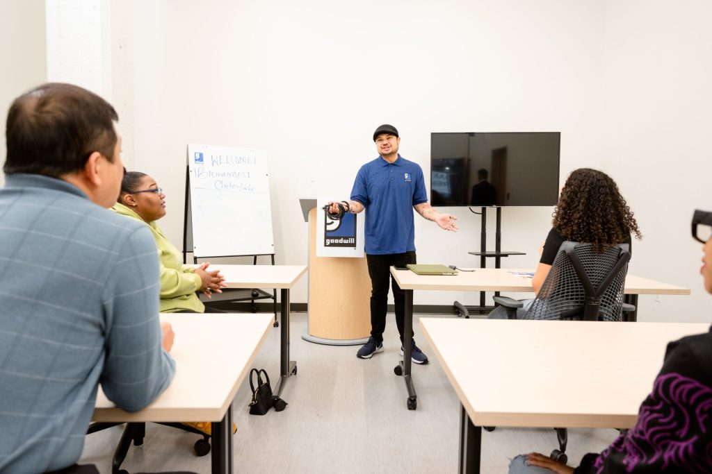 Presenter in blue polo and cap addresses a classroom audience with a whiteboard and a large TV behind.
