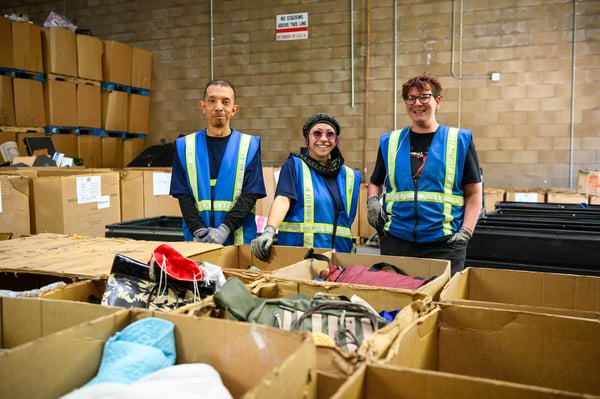 Three volunteers in safety vests standing behind open cardboard boxes filled with clothing in a warehouse.