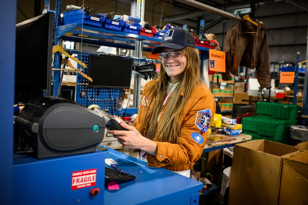 Young woman in an orange jacket and cap uses a handheld barcode scanner at an industrial workstation.