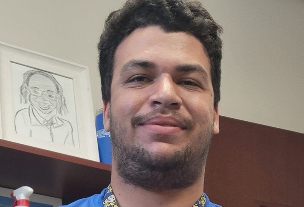 Smiling man taking a selfie in an office, with a framed pencil sketch on a shelf behind him.