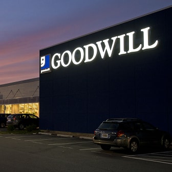 Exterior of a Goodwill thrift store at dusk with an illuminated sign and parked cars.