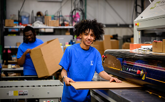 Young worker in a blue shirt operates a large industrial printer as another colleague moves a box in a busy workshop.