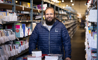 Smiling bearded man in a blue jacket stands in a bookstore aisle surrounded by shelves of books.