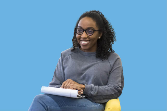 Smiling woman wearing glasses sits with a notebook on her lap against a blue background.