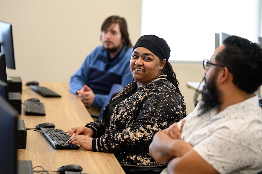 Smiling woman with a black head wrap sits at a computer in a classroom, with two colleagues nearby.