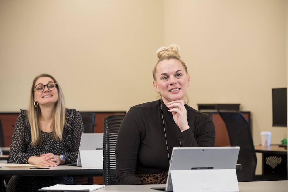 A woman in a black top with a blonde bun sits at a desk, thoughtful, while another smiling woman with glasses sits behind her.