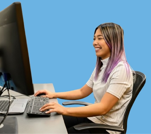 Person with purple-tinted hair using a desktop computer at a desk against a solid blue background.