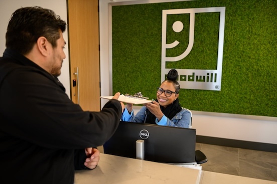 Smiling receptionist hands a plate of food to a customer at a modern reception desk with a green moss wall.