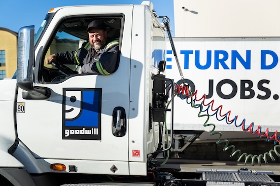 Truck driver sits in the cab of a white Goodwill donation truck, with a large Goodwill logo on the door and colorful hoses along the trailer.