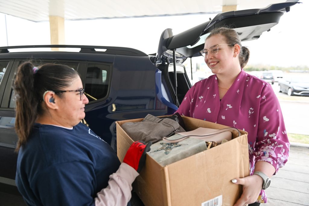 Two women unload a cardboard box from a hatchback during a drive-through donation, smiling at each other.