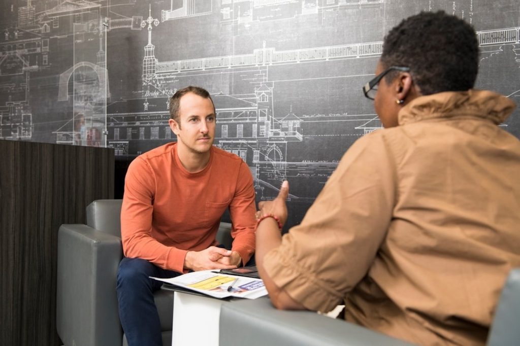 Man in orange shirt chats with another person across papers, in a lounge with a chalkboard wall of architectural sketches.