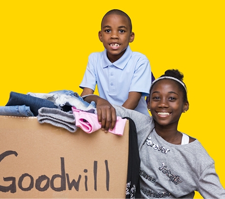 Two smiling children with a pile of donated clothes beside a Goodwill box against a bright yellow background.