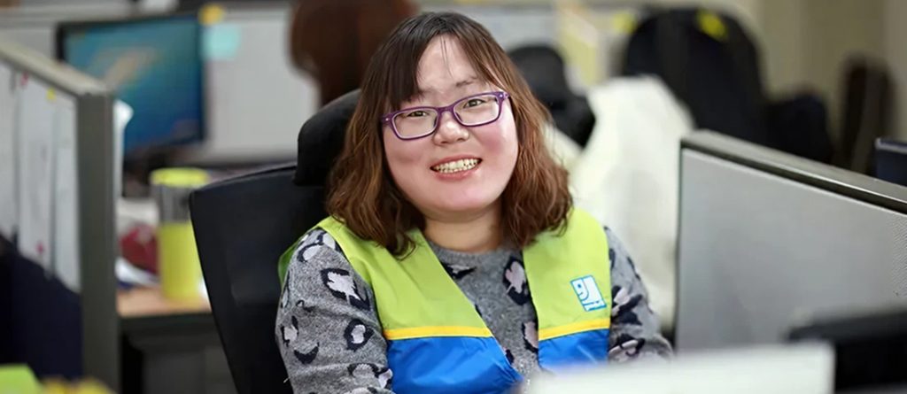 Smiling woman wearing a neon safety vest seated at a desk in a busy office.
