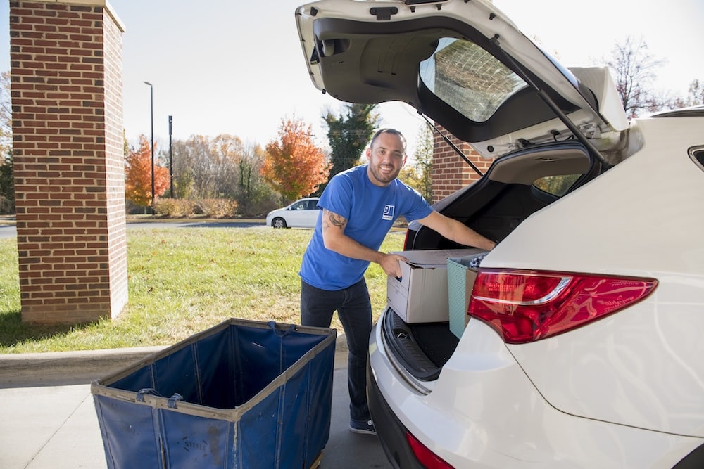 Delivery worker in a blue shirt loads boxes into the trunk of a white SUV in a parking lot.
