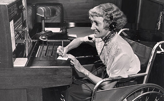 Woman in a wheelchair wearing a headset, writing at a vintage office desk with a telephone.