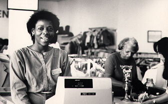 Smiling cashier with name badge at a checkout counter in a busy thrift shop.