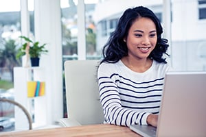 Smiling woman sits at a desk, working on a laptop in a bright, modern office.