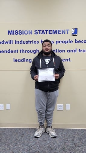 Man standing in front of a mission-statement wall holding a certificate.