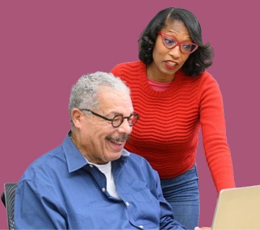 Smiling elderly man in blue shirt using a laptop, assisted by a woman in a red sweater.