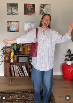 Woman standing with arms outstretched in a living room, smiling, wearing a striped shirt and jeans.