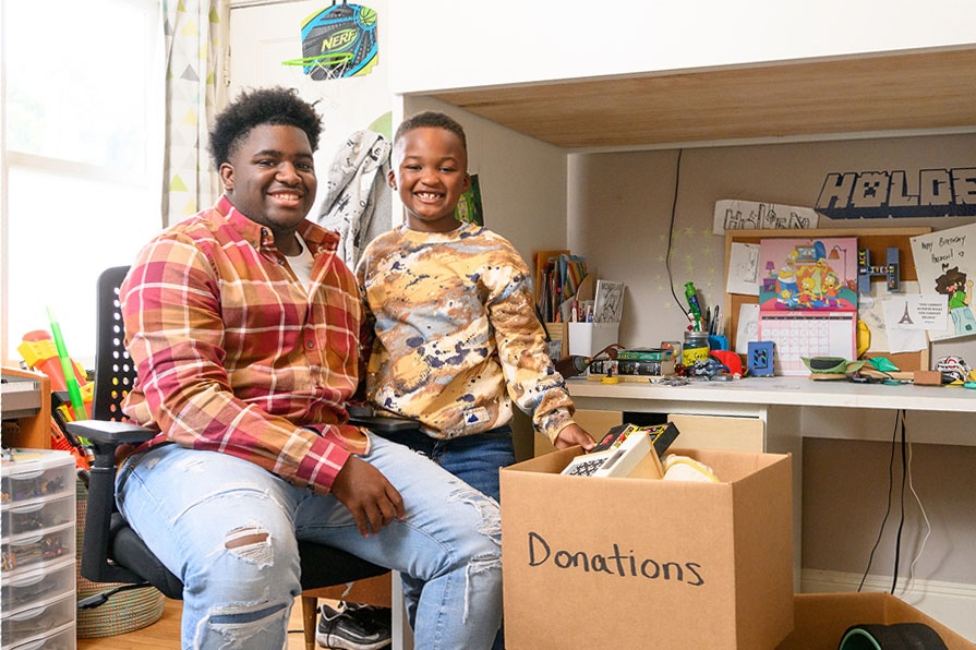 Two smiling people sit beside a cardboard box labeled Donations in a bright, cluttered room.