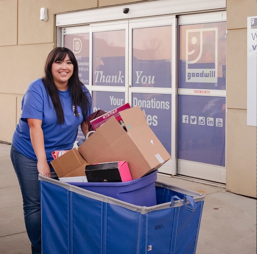 Volunteer woman in a blue shirt pushes a large blue cart filled with cardboard boxes toward a Goodwill donation entrance.