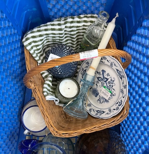 Basket of assorted household items, plate, glassware, candle, towel inside a blue crate.