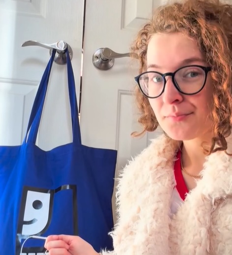 Young person with curly hair and glasses posing indoors beside a blue tote bag hanging on a door handle.