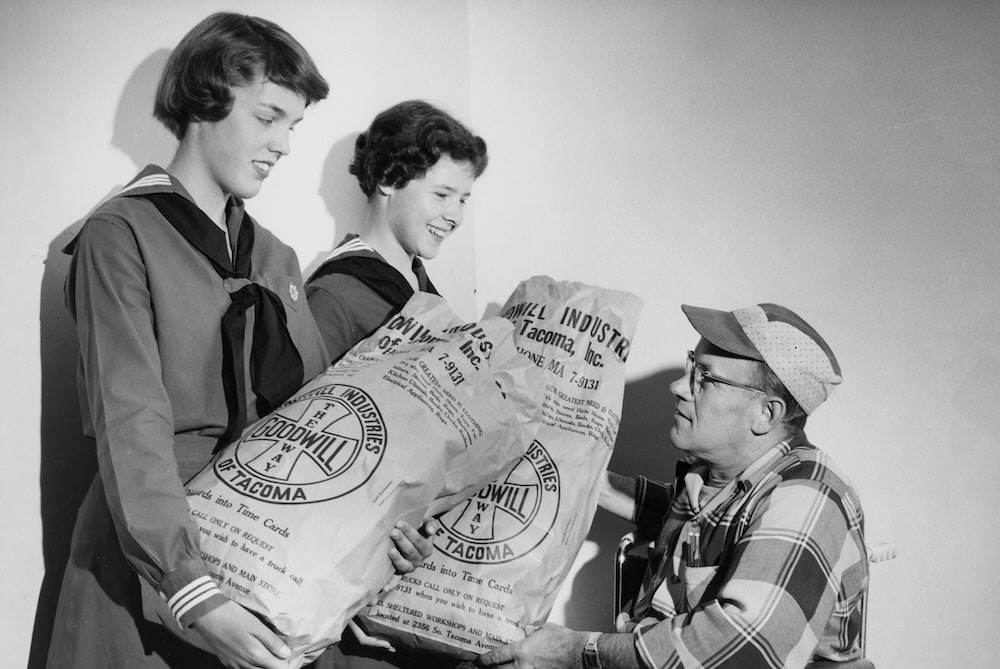Two young women in sailor uniforms hand a Goodwill donation bag to a man in a plaid shirt and visor.