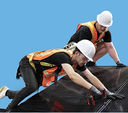 Two construction workers in hard hats and orange vests kneel on a sloped roof, securing black roofing material.