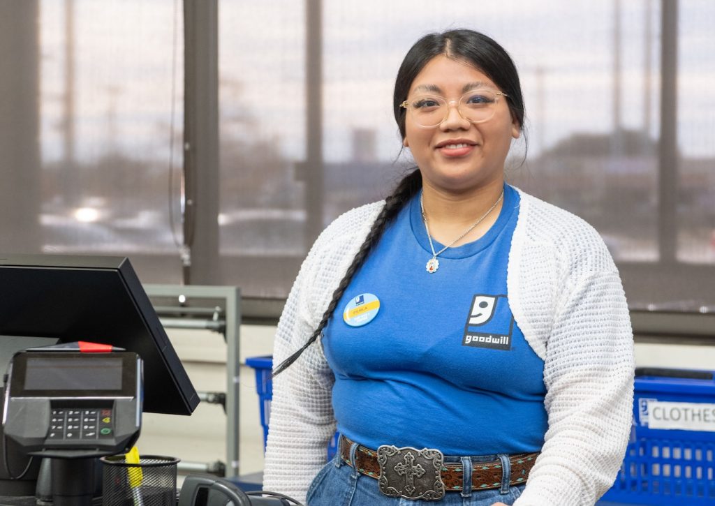 Smiling woman cashier at a Goodwill checkout wearing a blue shirt, white cardigan, and a decorative belt.