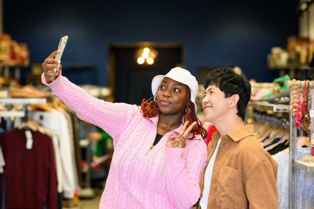 Two friends take a selfie in a clothing store; one wears a pink sweater and white bucket hat.