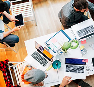 Group of people collaborating around a table with laptops, papers, and sticky notes in a bright office.