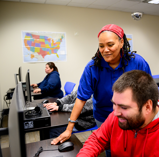 Instructor assists a student at a computer in a classroom, with another student working in the background and a United States map on the wall.