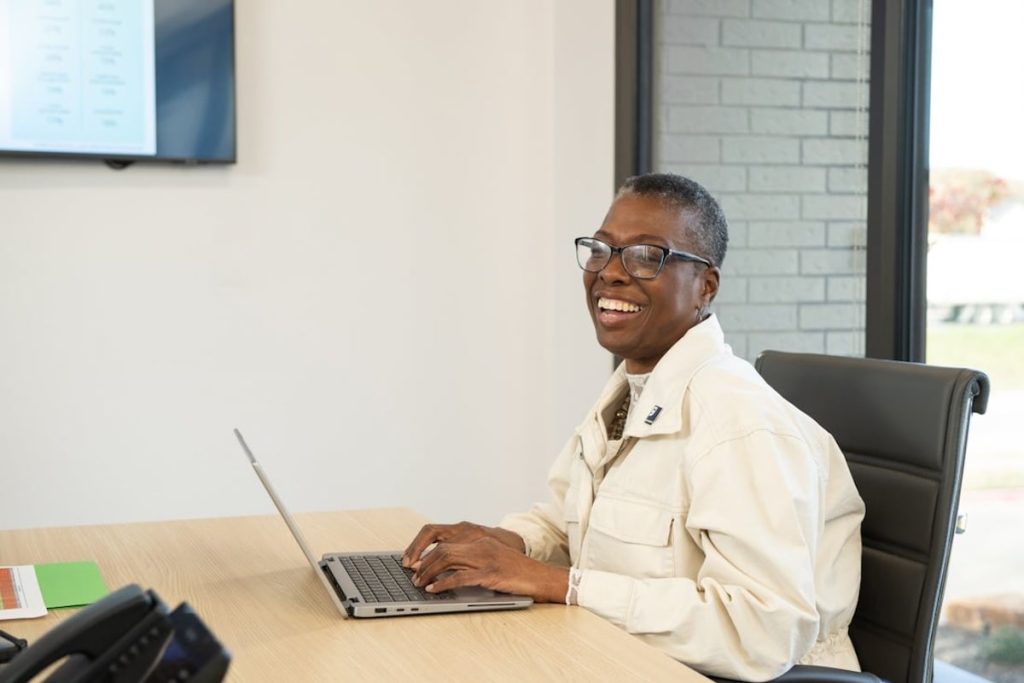 Smiling woman wearing a beige jacket and glasses typing on a laptop at a desk in a bright office.