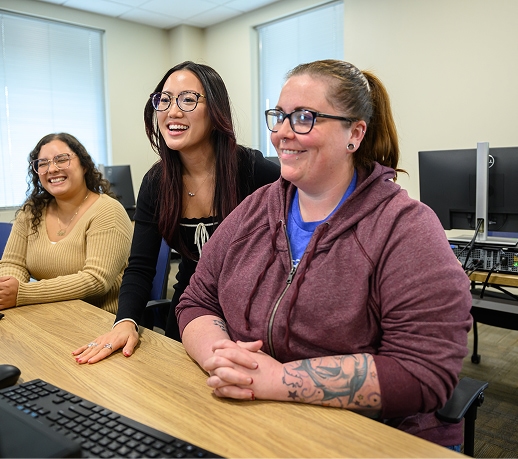 Three women sit at a wooden table in a computer lab, smiling as they look toward a screen.