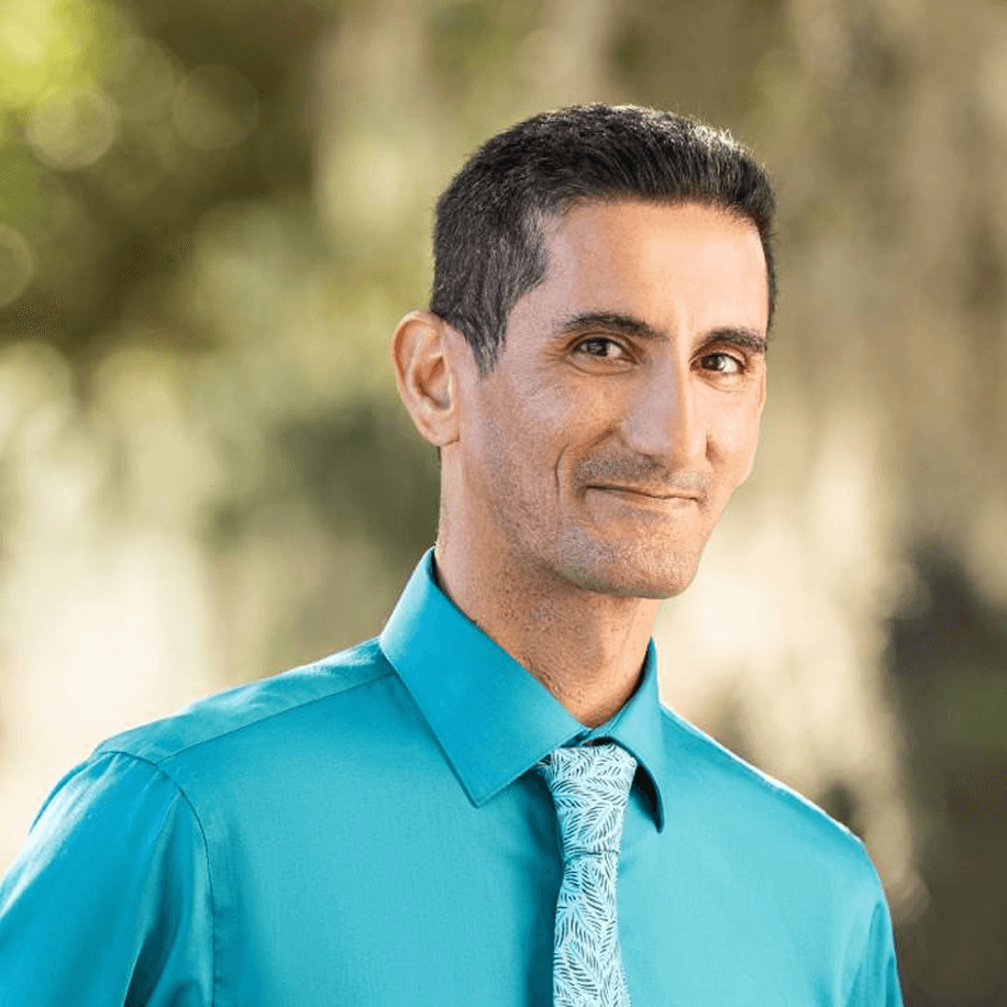 Smiling man in turquoise shirt and patterned tie outdoors.