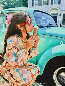 Women wearing red sunglasses and a colorful jumpsuit standing in front of a blue car eating an ice cream cone
